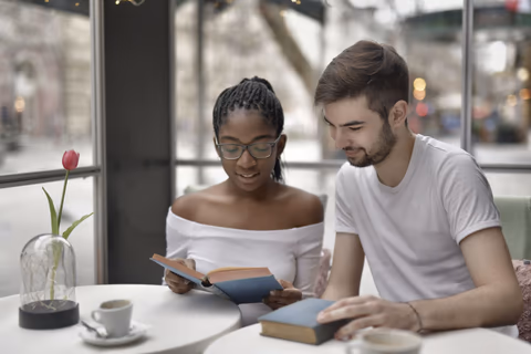 Dos personas leen juntas en una cafetería, con libros y tazas de café sobre la mesa